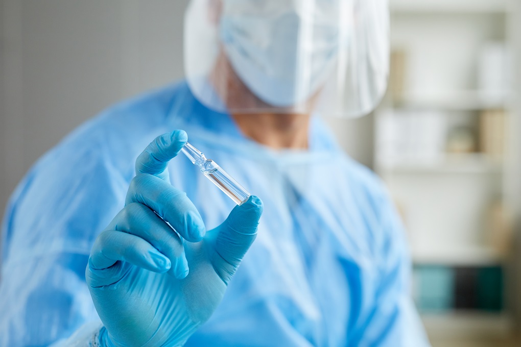 Head cropped close-up shot of unrecognizable medical worker wearing personal protective uniform holding ampoule with vaccine